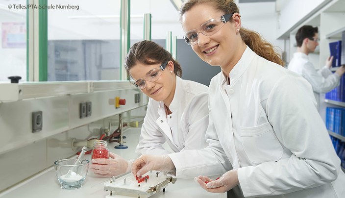 Zwei Pharmazeutisch-technische Assistent:innen (PTA) bei Ihrer Arbeit im Labor in Kitteln und mit Schutzbrillen Zwei Pharmazeutisch-technische Assistent:innen (PTA) bei Ihrer Arbeit im Labor in Kitteln und mit Schutzbrillen
