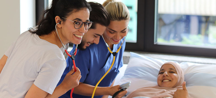 Drei junge lächelnde Personen in Pflegebekleidung mit Stethoskop am Bett einer Patientin, die Daumen hoch zeigt Drei junge lächelnde Personen in Pflegebekleidung mit Stethoskop am Bett einer Patientin, die Daumen hoch zeigt