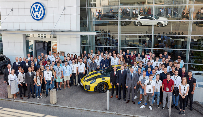Eine große Gruppe steht vor einem VW-Gebäude um ein gelb-schwarzes Sportauto herum. Alle blicken zur Kamera.