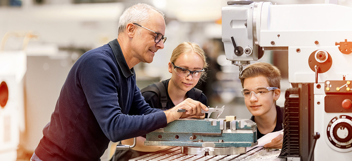 Drei Personen stehen an einer großen Maschine in einer Werkstatt. Eine Person erklärt etwas, die anderen hören aufmerksam zu. Alle tragen Schutzbrillen. Drei Personen stehen an einer großen Maschine in einer Werkstatt. Eine Person erklärt etwas, die anderen hören aufmerksam zu. Alle tragen Schutzbrillen.
