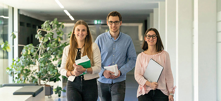 Drei Personen gehen einen Gang entlag und halten Akten oder Bücher in den Händen. Drei Personen gehen einen Gang entlag und halten Akten oder Bücher in den Händen.